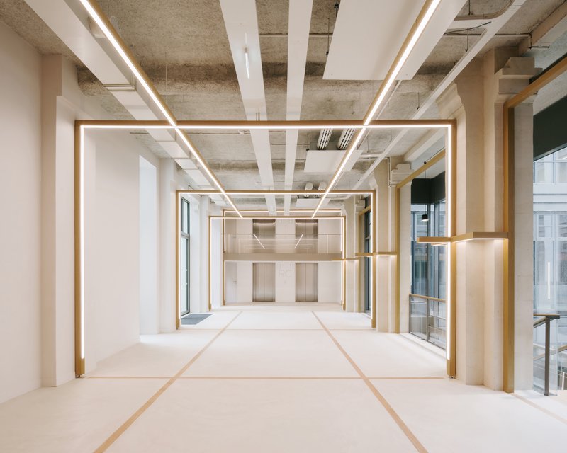 Open floor space with repeated brass-trimmed light portals beneath exposed concrete ceiling and white beam inserts
