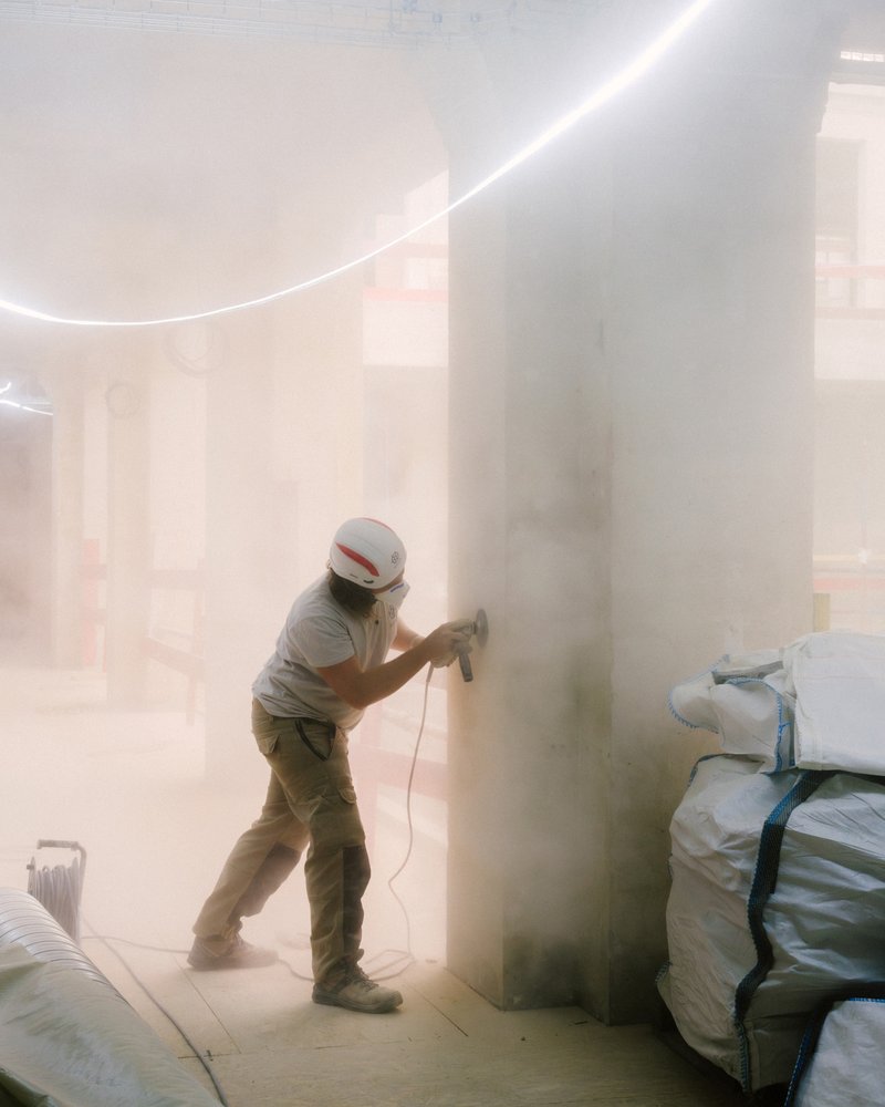 Worker sanding a cylindrical column in a dust-filled interior with curved cove lighting