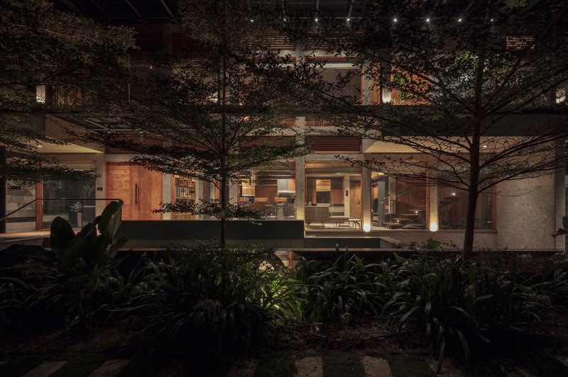 Night view of illuminated interior spaces behind floor-to-ceiling glass with mature trees in planted courtyard