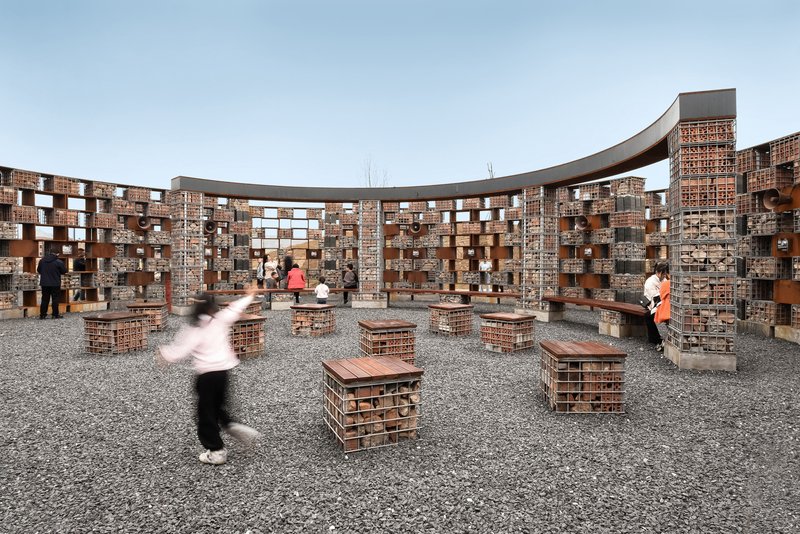 Courtyard with gravel ground and brick seating cubes enclosed by perforated brick and corten steel walls as visitors gather