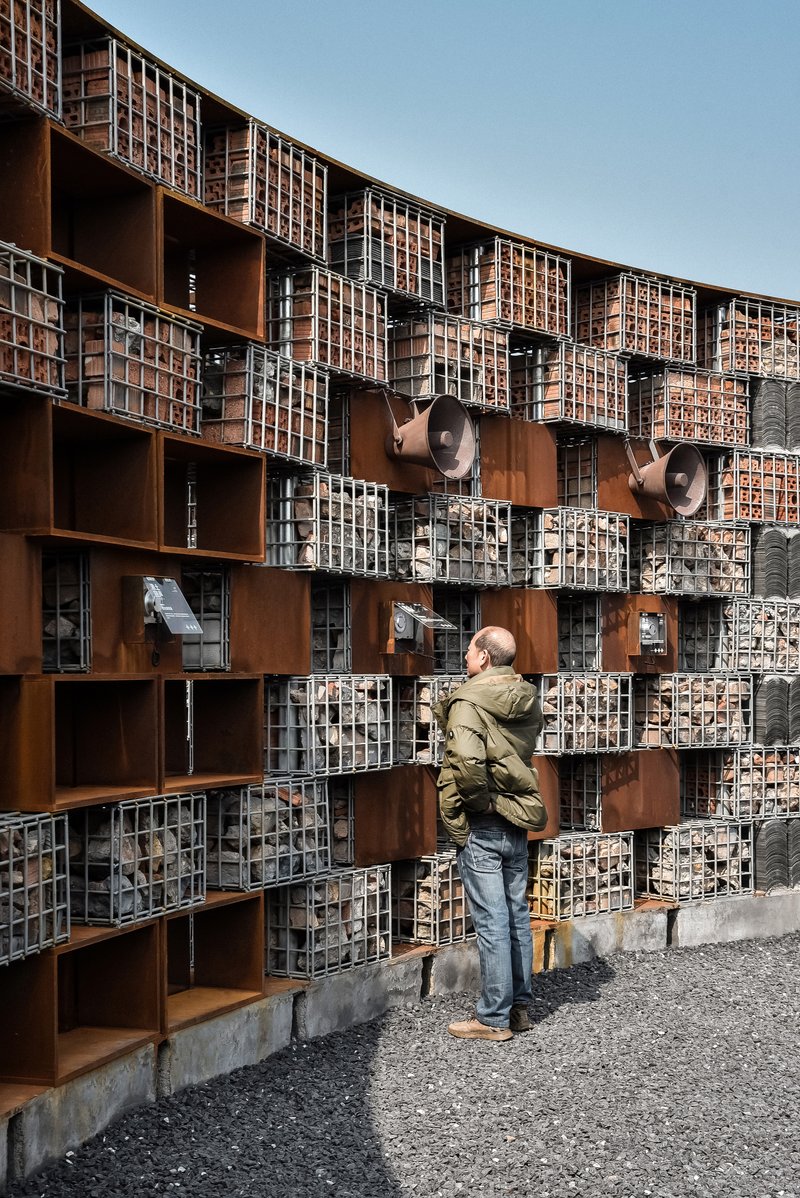 Visitor photographing the curved facade composed of wire-filled gabion baskets, corten frames and mounted speaker horns