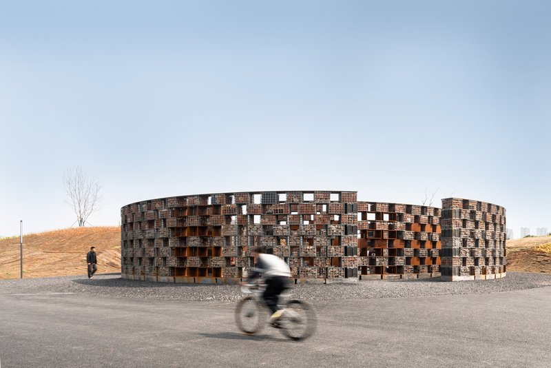 Cylindrical brick pavilion with a patterned facade beside a gravel path as a cyclist passes