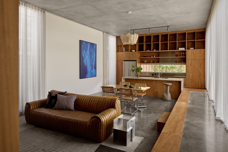 Open-plan living space with exposed concrete ceiling and timber joinery wall integrating kitchen and storage