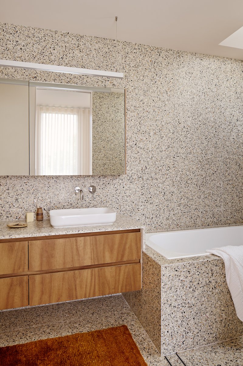 Bathroom interior with terrazzo walls, timber vanity, white vessel sink, and recessed bathtub