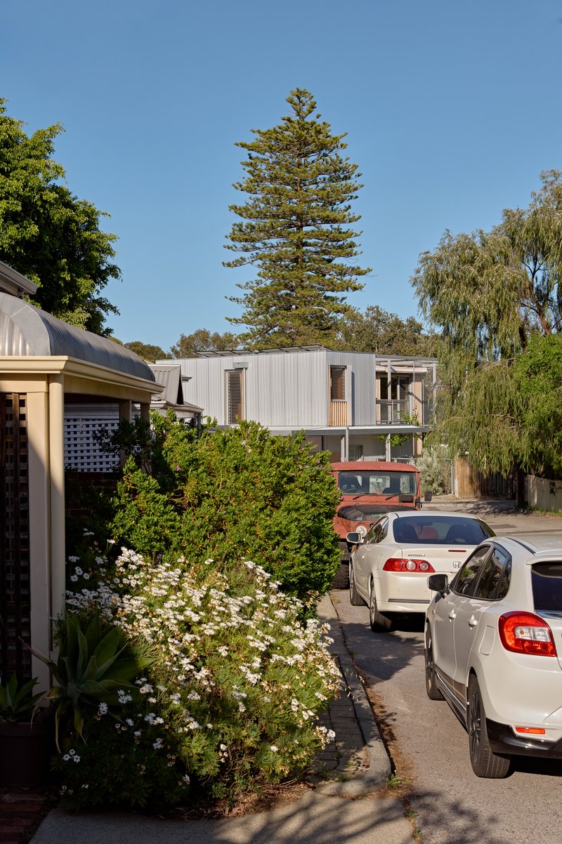Street view of corrugated metal volume elevated above parked cars and mature trees