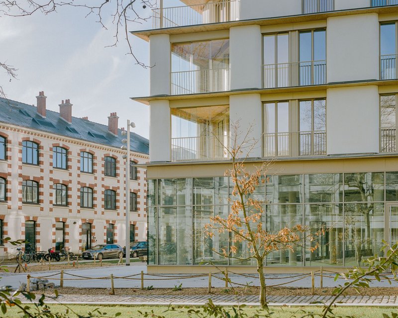Ground floor glass curtain wall beneath upper residential levels with cylindrical columns and bare tree