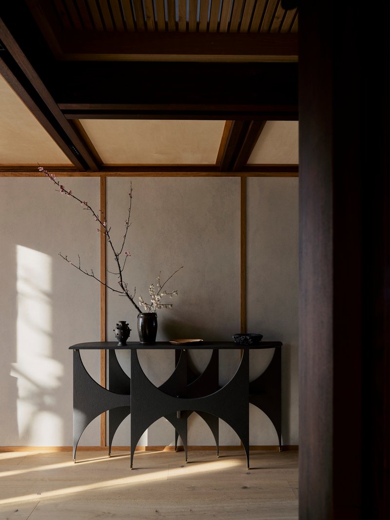 Sculptural black console table with flowering branch against timber-framed wall in morning light