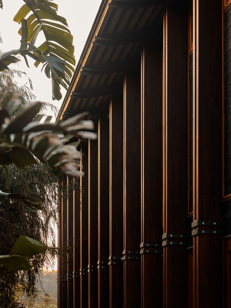 Vertical timber battens on dark facade with metal connectors beneath deep eaves and tropical foliage