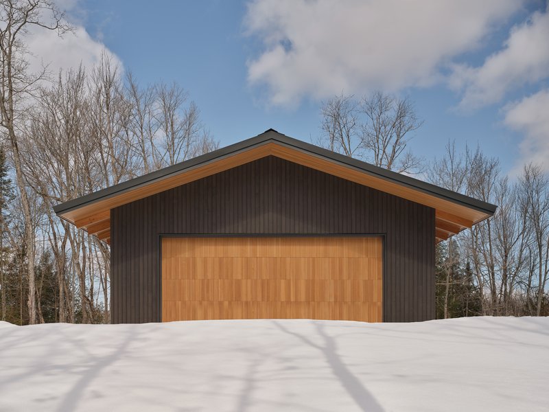 Front elevation showing timber garage door and extended roof overhangs casting shadows on fresh snow