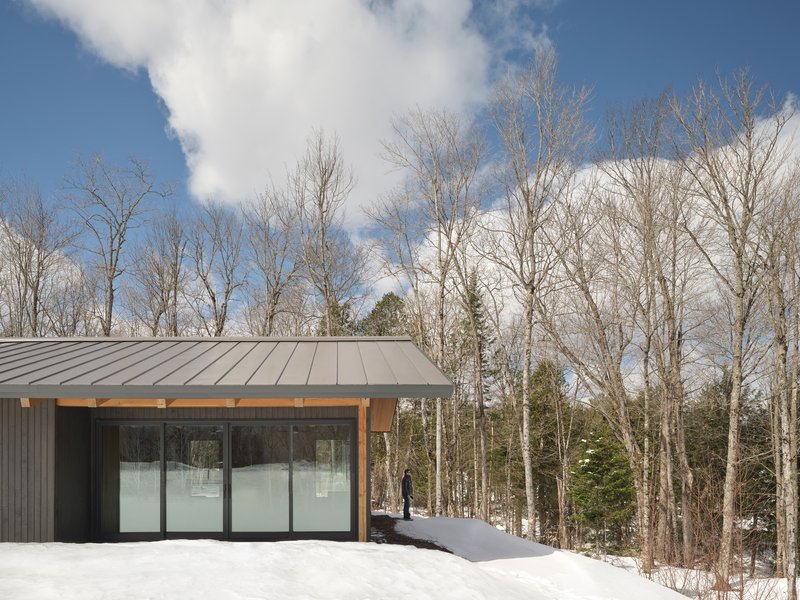 Dark vertical siding and glass doors of the volume beneath a standing seam metal roof in snow