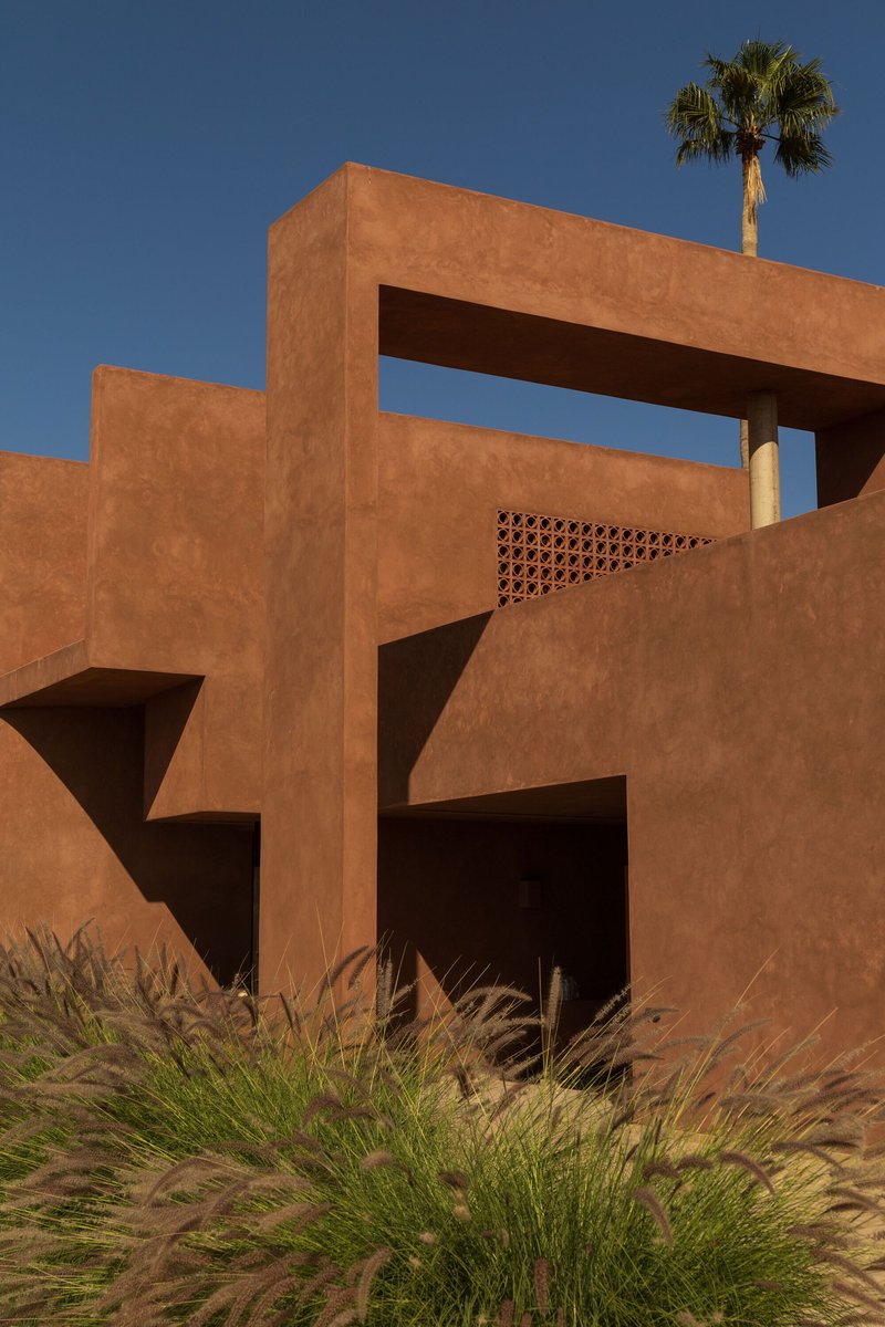 Layered terracotta-colored stucco volumes with perforated screen panel and palm tree rising behind ornamental grasses