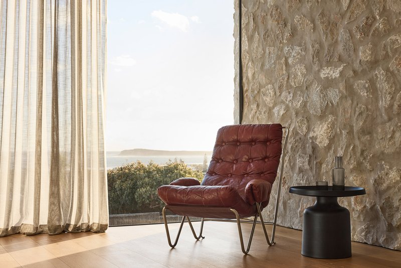 Interior corner with textured stone wall, sheer curtains, and leather armchair overlooking a coastal view