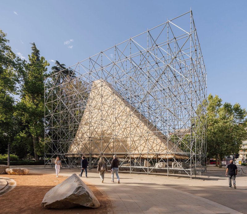 Triangular timber pavilion enclosed by a steel scaffolding framework in a public plaza with scattered stones