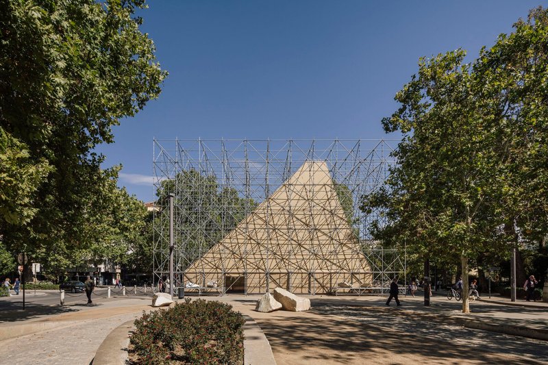 Frontal view of the scaffolding structure framing the timber gable form between tree-lined pathways