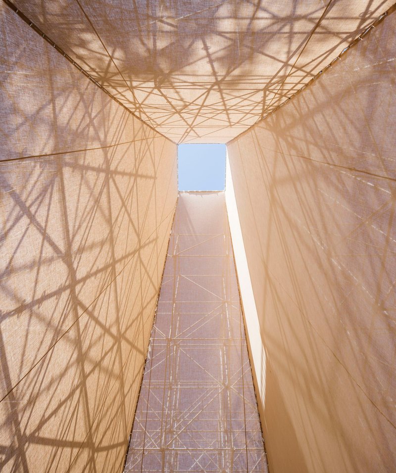 Upward view through the central void showing steel trusses and scaffold shadows on translucent fabric surfaces