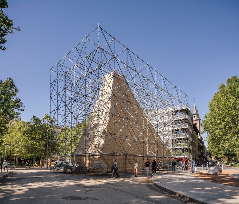 Triangulated steel scaffolding structure enclosing a gabled volume in a tree-lined plaza with pedestrians