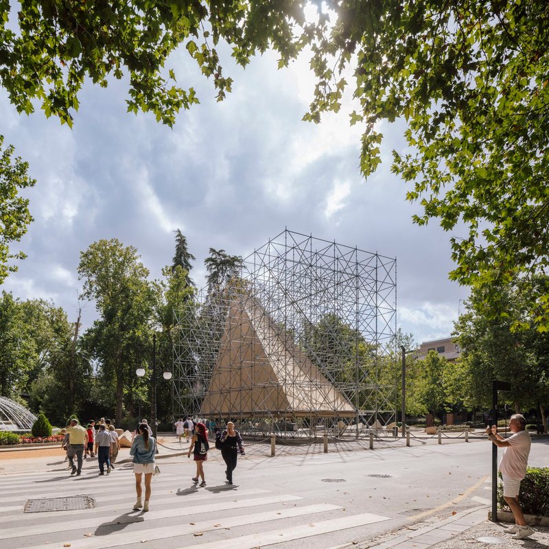 Scaffolding pavilion with burlap panels in a park plaza with visitors and overhanging tree branches