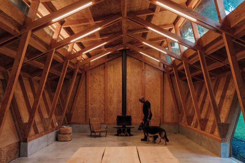 Main hall with a person and dog beside a central stove under the pitched timber frame at dusk
