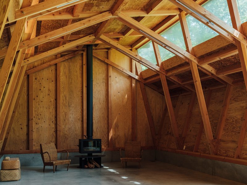Plywood-clad interior wall with a wood stove on the concrete floor beneath angled timber trusses
