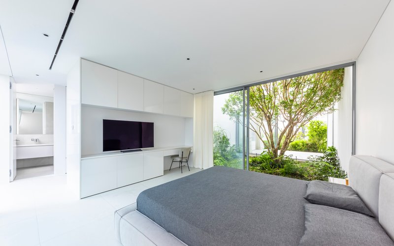 White bedroom with floor-to-ceiling glazing opening onto a courtyard garden with a mature tree