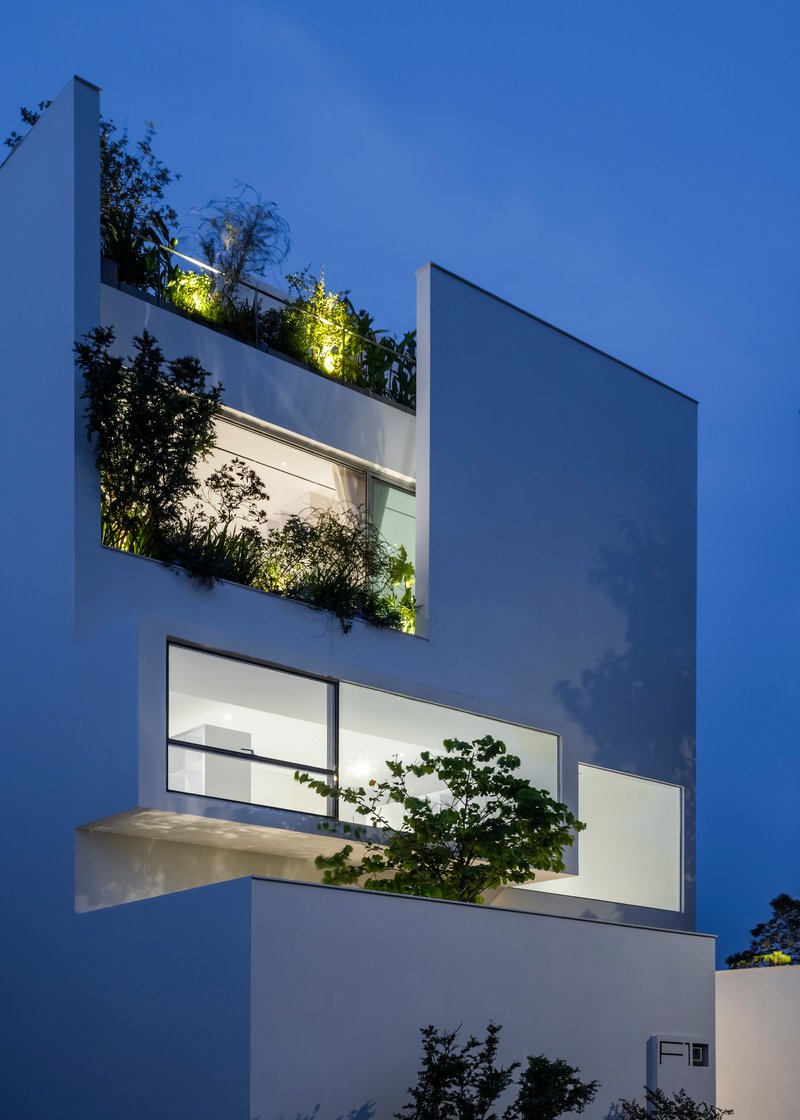 White facade with recessed planted terraces and illuminated greenery against a darkening evening sky