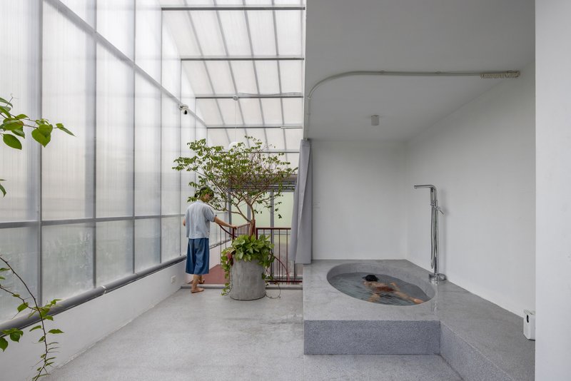 Interior bath terrace with raised concrete soaking tub and person standing near potted tree by translucent window wall