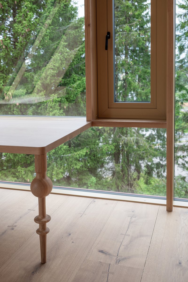 Turned timber balustrade detail with spherical joint beside floor-to-ceiling window overlooking forest