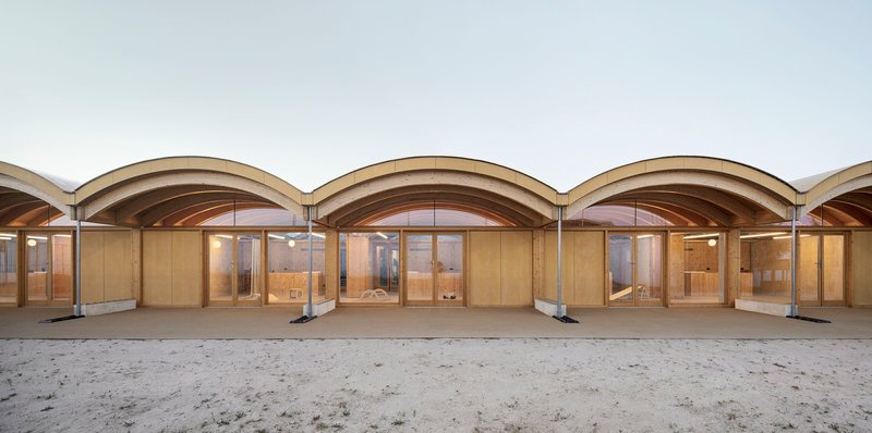 Covered colonnade with repeated arched timber vaults and glazed timber doors opening onto a sandy courtyard