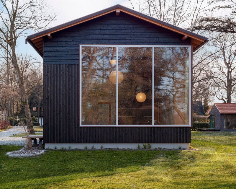 Charred timber gable facade with large tri-panel window reflecting bare trees in late afternoon light