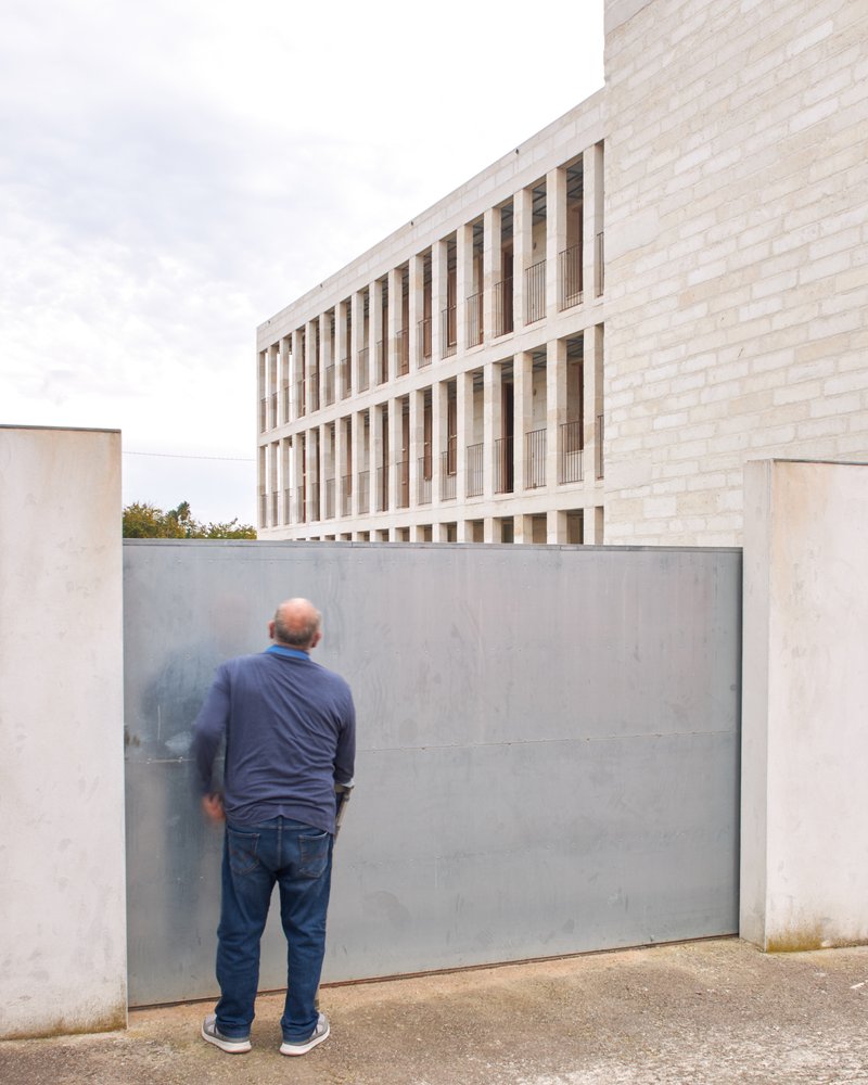 Figure standing at a steel gate below the gridded stone facade in soft light