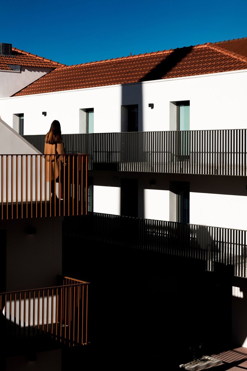 Timber-railed balconies and dark metal screens beneath terracotta tile roofs with figure in afternoon shadow