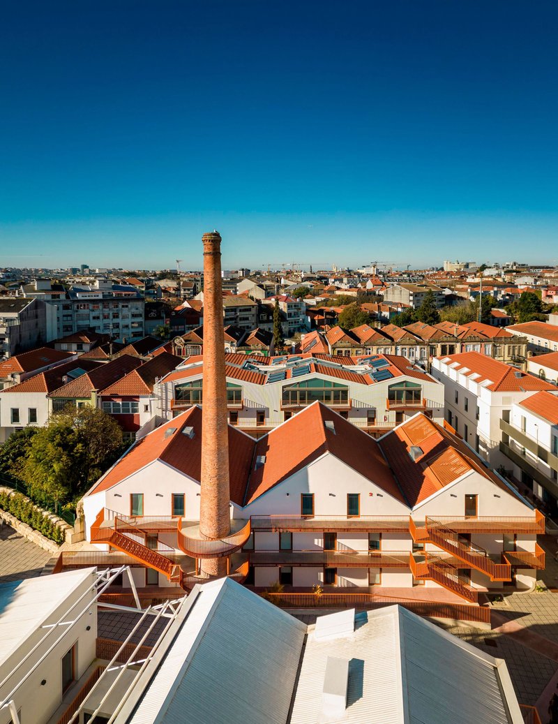 Rooftop panorama of gabled volumes with terracotta tiles and a cylindrical brick chimney under clear sky