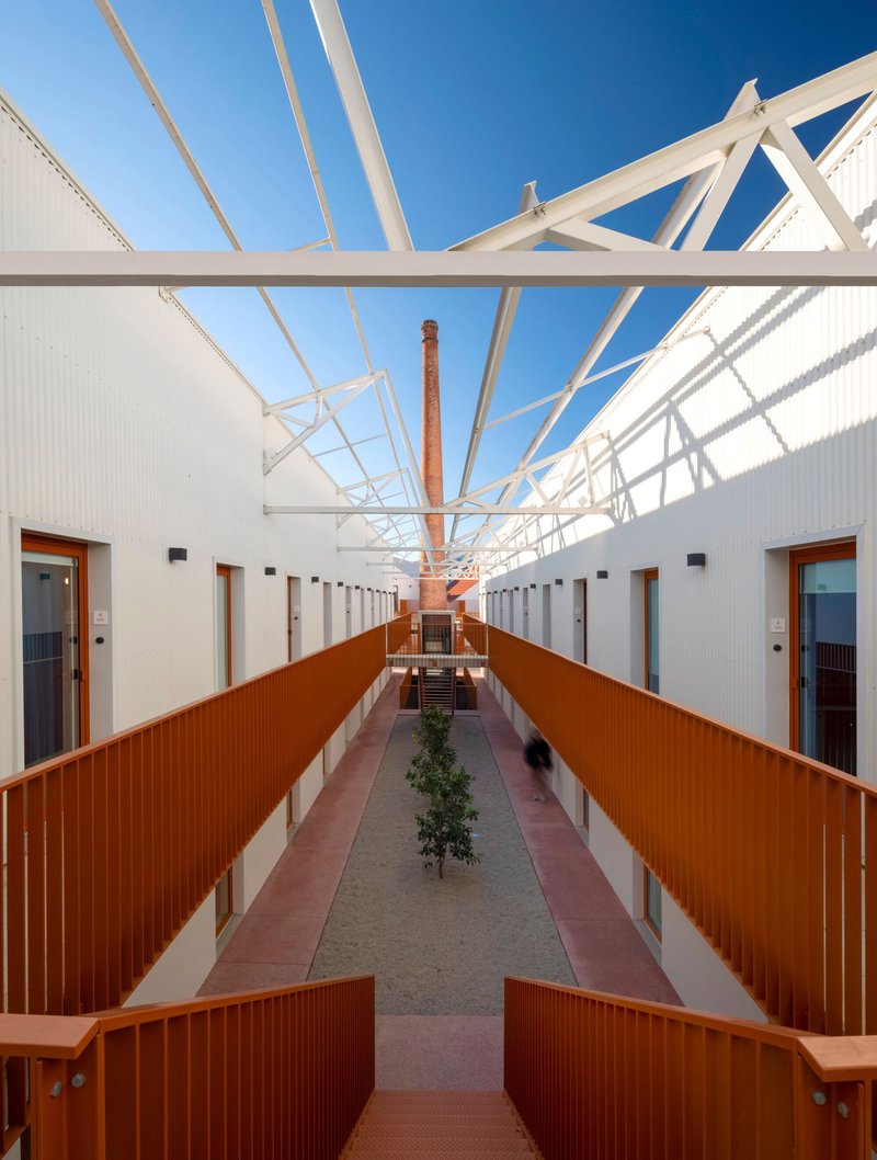 Interior corridor with exposed white timber trusses, terracotta metal railings, and a planted tree