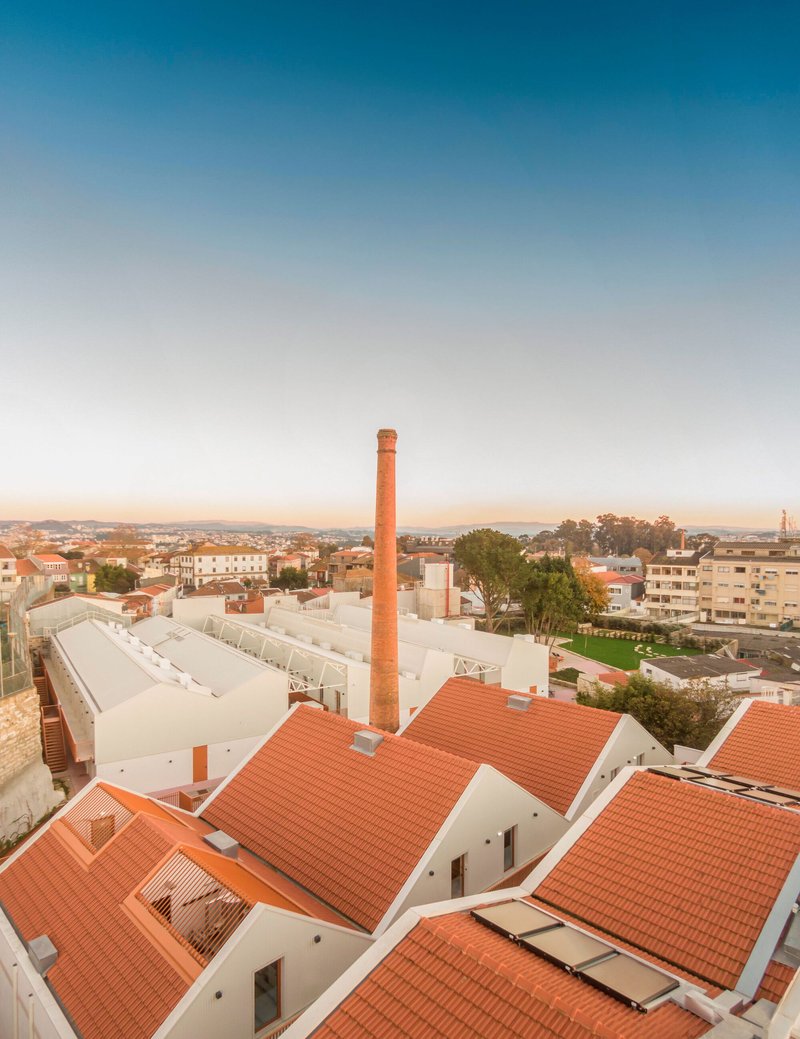 Roofscape with industrial chimney and terracotta-tiled volumes set against low-rise residential surroundings at dusk