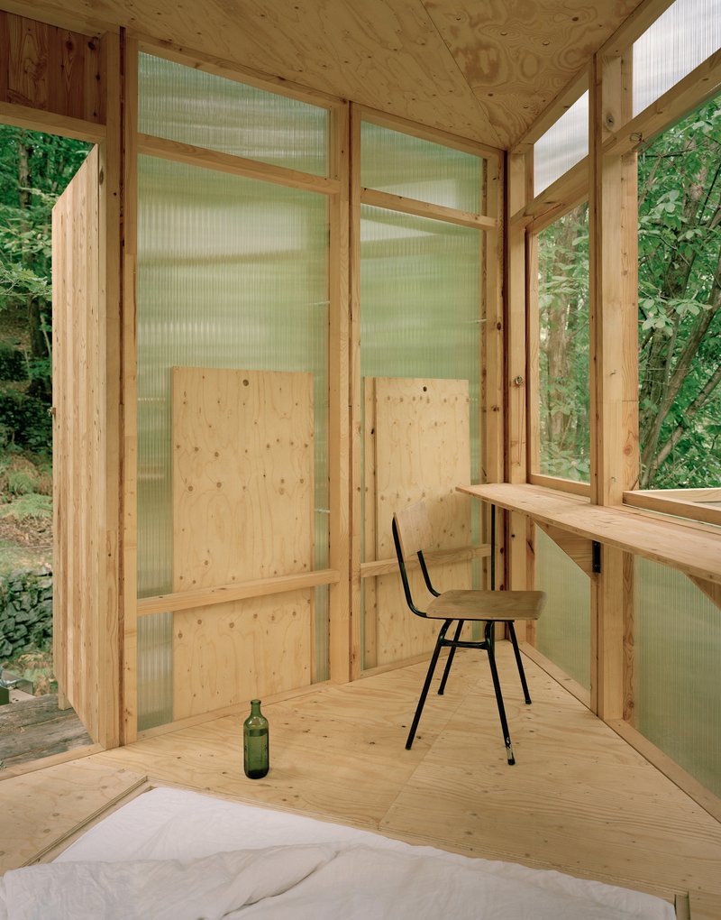 Interior corner with plywood walls, translucent panel partitions, folding desk, chair, and bedding on floor