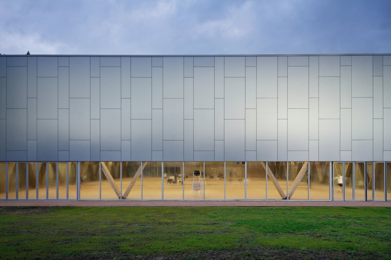 Long facade with pale panel cladding over timber diagonal bracing visible through full-height glazing at dusk