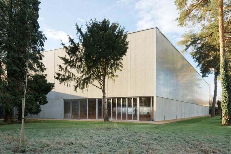 Corner view of the pale metal-clad hall beside mature conifers on frost-covered grass at dawn