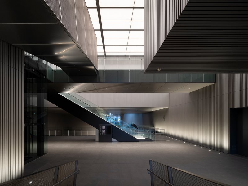 Ground floor lobby with cantilevered stair platform hovering above under a gridded skylight