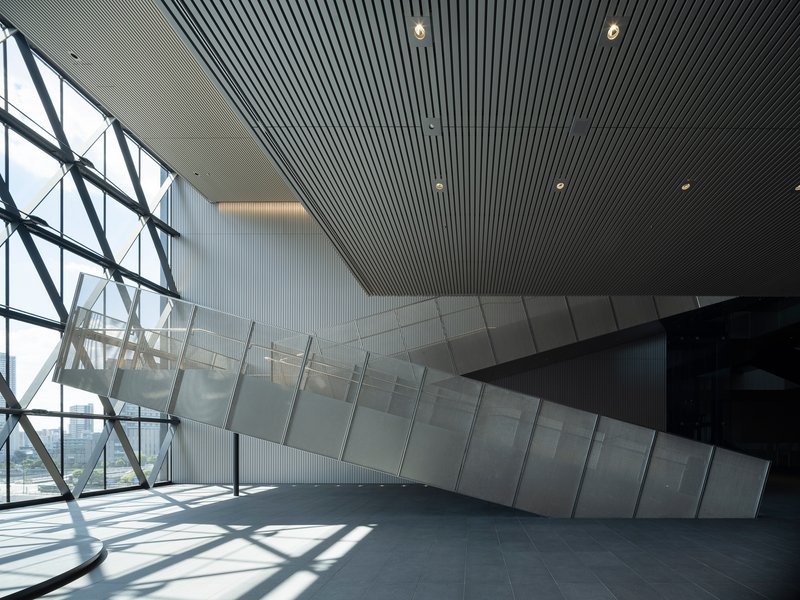 Diagonal steel bracing meets glass and ribbed ceiling panels with sunlight casting sharp shadows across the floor