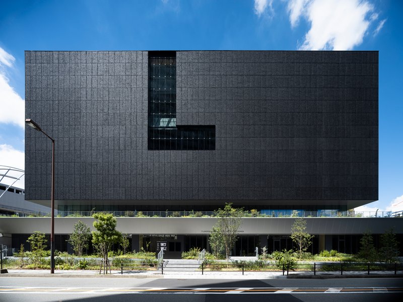 Street view of the dark tiled facade elevated above a landscaped plaza with young trees