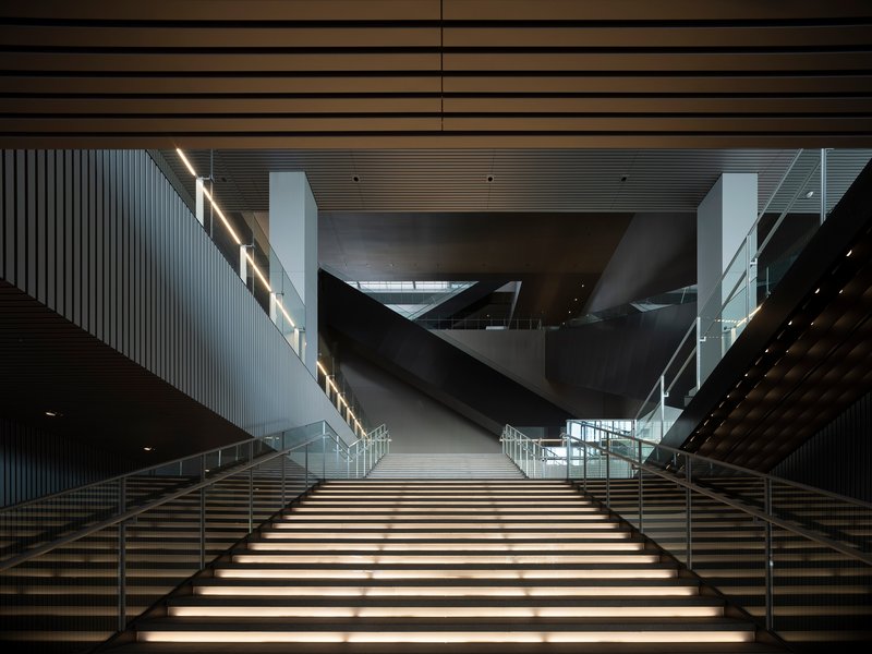 Wide staircase with integrated lighting rising toward angular volumes suspended in the atrium