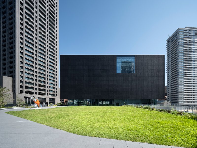 Dark textured facade with single recessed window facing a lawn and residential towers beyond