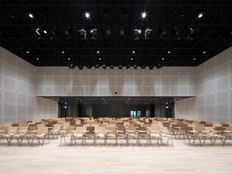 Performance hall with exposed rigging, white panel walls and wooden seating on pale flooring