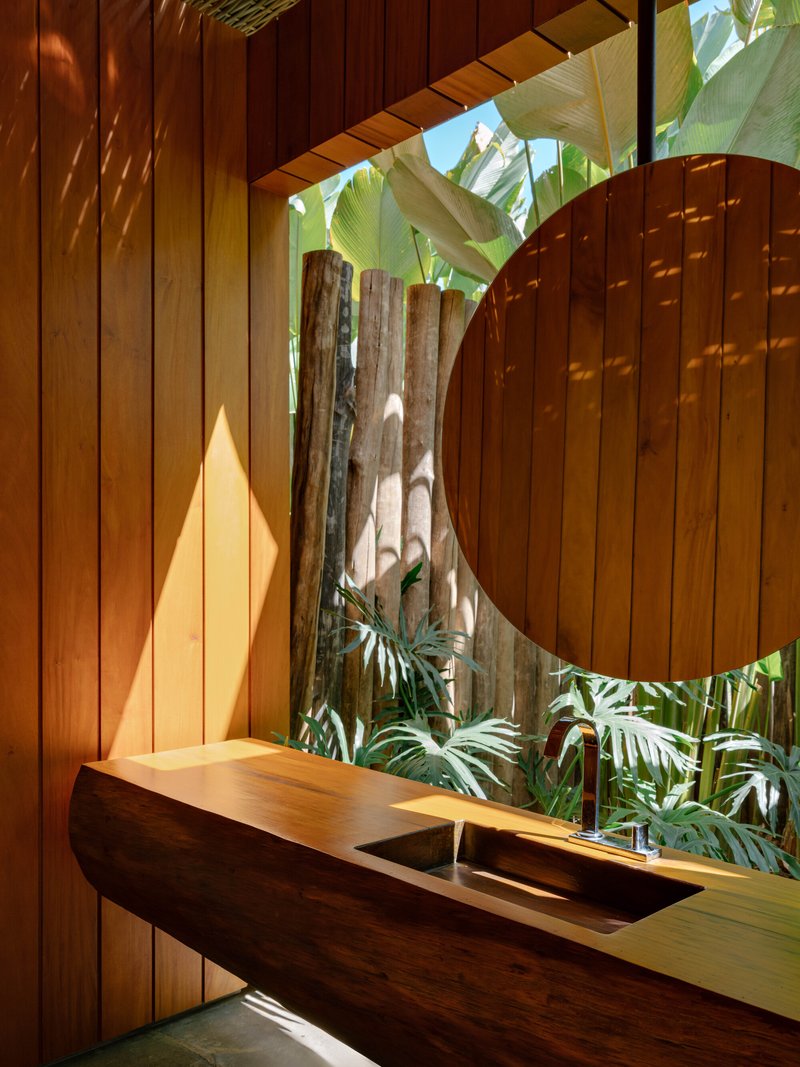 Timber vanity counter beneath a circular cutout in vertical wood slat wall with tropical plants beyond