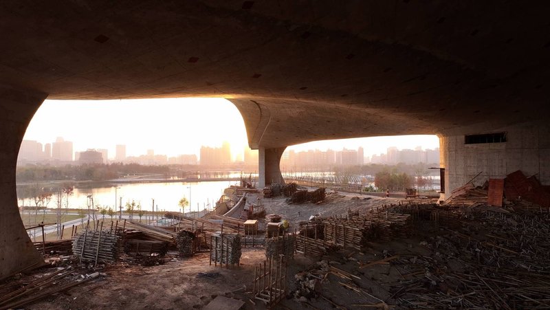 Arched openings in raw concrete shell framing a waterfront skyline at sunrise during construction