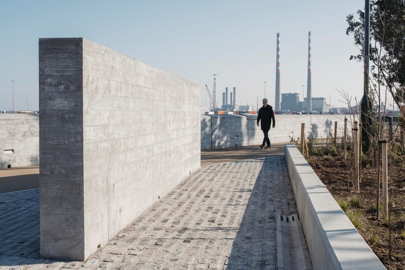 Cobblestone pathway along concrete parapet wall with pedestrian and industrial stacks in distance
