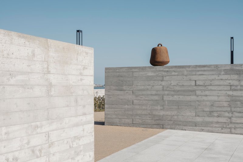 Courtyard framed by board-formed concrete walls with weathered bell suspended from upper parapet