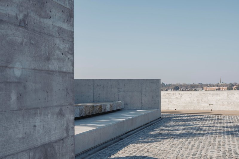 Granite bench built into curved concrete wall overlooking cobblestone plaza and distant shoreline