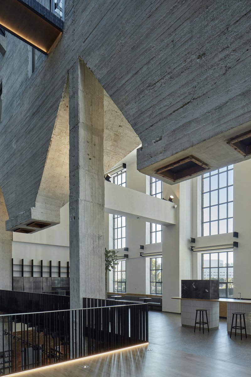 Angled concrete column supporting an elevated slab above a communal kitchen with stools and potted plant