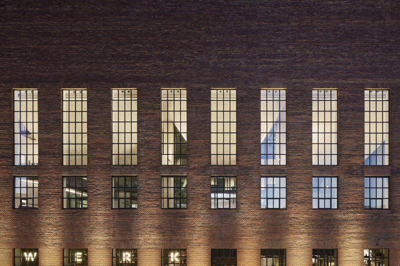 Dark brick facade with tall gridded windows illuminated from within at dusk
